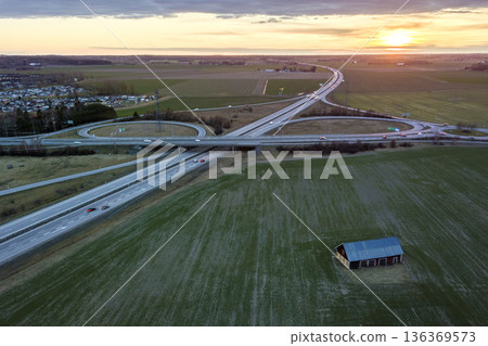 Aerial view of wooden barn in green field on background of modern highway road intersection at dawn on sunny spring day. Drone photography. Aerial view of wooden barn in green field on background of modern highway road intersection at dawn on sunny spring day. Drone photography. 136369573