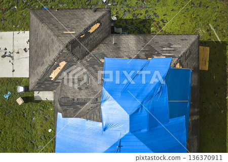 Aerial view of damaged in hurricane Ian house roof covered with blue protective tarp against rain water leaking until replacement of asphalt shingles Aerial view of damaged in hurricane Ian house roof covered with blue protective tarp against rain water leaking until replacement of asphalt shingles 136370911