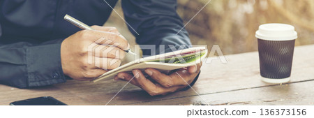 Man hands writing notebook diary with coffee cup and smartphone on wood desk. Close up man hands using pen sitting at wooden table outdoors lifestyle. Men write planner note diary office agenda Man hands writing notebook diary with coffee cup and smartphone on wood desk. Close up man hands using pen sitting at wooden table outdoors lifestyle. Men write planner note diary office agenda 136373156