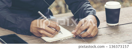 Man hands writing notebook diary with coffee cup and smartphone on wood desk. Close up man hands using pen sitting at wooden table outdoors lifestyle. Men write planner note diary office agenda Man hands writing notebook diary with coffee cup and smartphone on wood desk. Close up man hands using pen sitting at wooden table outdoors lifestyle. Men write planner note diary office agenda 136373157