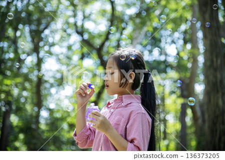 Adorable happy cute girl playful foam bubbles in green playground in summer outdoors. Funny Cheerful girl in the park with happy times. Adorable happy cute girl playful foam bubbles in green playground in summer outdoors. Funny Cheerful girl in the park with happy times. 136373205