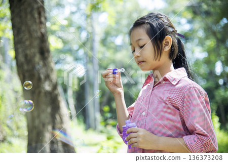 Adorable happy cute girl playful foam bubbles in green playground in summer outdoors. Funny Cheerful girl in the park with happy times. 136373208