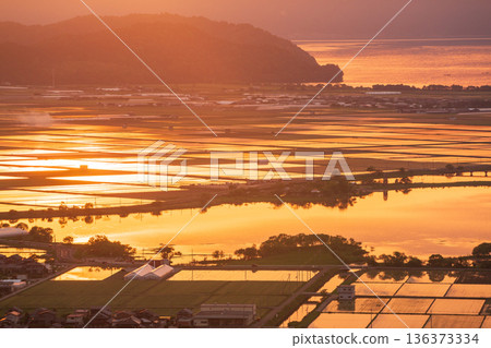 從滋賀縣東近江市豬子町的豬子山上眺望，傍晚時分，稻田在播種前的夕陽映照下泛著橙紅的光芒。 136373334
