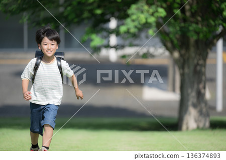 A boy (boy) running with a school bag on his back in the schoolyard with the school building in the background, looking at the camera with a smile A boy (boy) running with a school bag on his back in the schoolyard with the school building in the background, looking at the camera with a smile 136374893