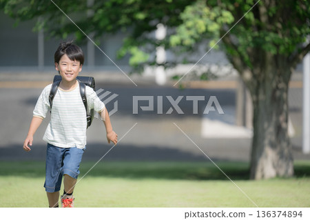 一個男孩背著書包在操場上奔跑，背景是學校教學大樓，他微笑著看著鏡頭。 136374894