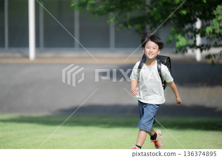 A boy (boy) running with a school bag on his back in the schoolyard with the school building in the background, looking at the camera with a smile A boy (boy) running with a school bag on his back in the schoolyard with the school building in the background, looking at the camera with a smile 136374895