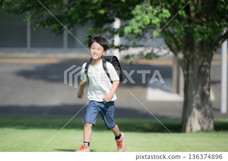 A boy (boy) running with a school bag on his back in the schoolyard with the school building in the background, looking at the camera with a smile A boy (boy) running with a school bag on his back in the schoolyard with the school building in the background, looking at the camera with a smile 136374896