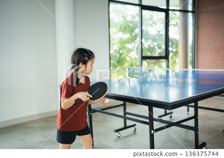 Young girl playing table tennis in bright room with large windows, showcasing focus and determination in recreational sport activity Young girl playing table tennis in bright room with large windows, showcasing focus and determination in recreational sport activity 136375744