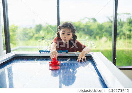 Young girl playing air hockey indoors with concentration and excitement, natural light streaming through large windows, green landscape in background Young girl playing air hockey indoors with concentration and excitement, natural light streaming through large windows, green landscape in background 136375745