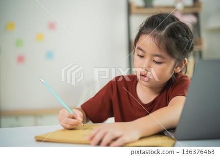 Young girl focused on drawing at a desk with laptop in a bright room filled with notes, representing creativity and education in home learning environment. Young girl focused on drawing at a desk with laptop in a bright room filled with notes, representing creativity and education in home learning environment. 136376463