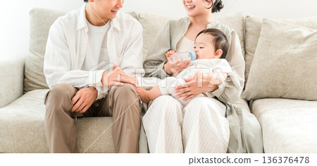 媽媽，一家人坐在家裡的沙發上，用奶瓶餵寶寶吃水 136376478