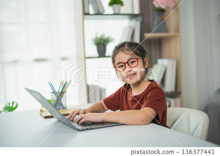 Young girl with glasses smiles while using laptop at home desk, surrounded by plants and bright window light, showcasing modern workspace creativity Young girl with glasses smiles while using laptop at home desk, surrounded by plants and bright window light, showcasing modern workspace creativity 136377441