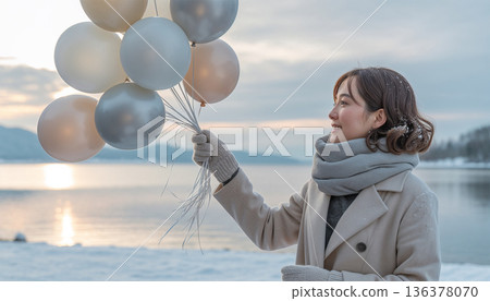 一位女子在白雪皚皚的湖岸邊舉起一個銀色氣球，表達著寧靜的冬日慶祝。 136378070