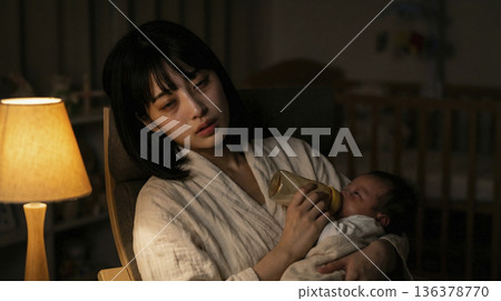 A mother with a blank expression is feeding her baby with a bottle in a dark room late at night 136378770