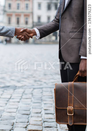 Two businessmen in suits shake hands outdoors on a cobblestone street, one holding a leather briefcase Two businessmen in suits shake hands outdoors on a cobblestone street, one holding a leather briefcase 136379223