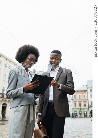 Two professionals, a woman and a man, are discussing documents on a clipboard while standing in an urban square 136379227