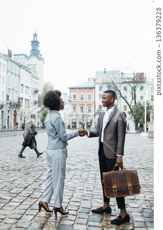 Two professionals, a man and a woman, shake hands on a cobblestone street in front of historic buildings 136379228