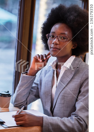 A professional Black woman with an afro hairstyle and glasses sits at a table near a window, taking notes on a clipboard while holding a pen and a coffee cup 136379350