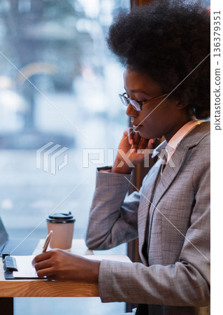 A focused woman in a suit and glasses writes on a clipboard in a cafe with a coffee cup nearby 136379351