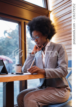 A focused Black woman in a suit writes in a notebook at a table in a cafe, with a tablet and coffee cup nearby A focused Black woman in a suit writes in a notebook at a table in a cafe, with a tablet and coffee cup nearby 136379352