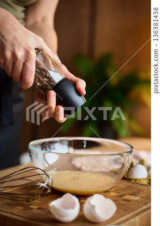 Hands grinding pepper into egg mixture in glass bowl on kitchen counter 136381438