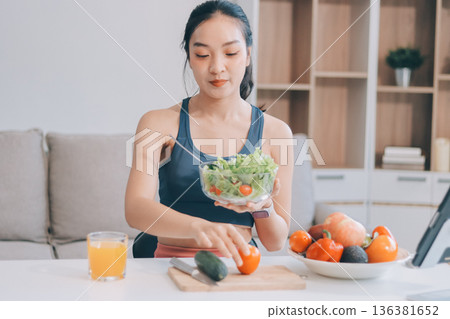 Fit young Asian women in activewear sitting on yoga mat and eating fresh vegetable salad after workout, representing clean eating, fitness lifestyle, wellness routine and health balance. 136381652