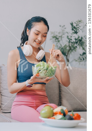 Fit young Asian women in activewear sitting on yoga mat and eating fresh vegetable salad after workout, representing clean eating, fitness lifestyle, wellness routine and health balance. 136381679