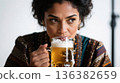 A young woman with dark hair sips a foamy beer from a glass mug, looking to the side. 136382659