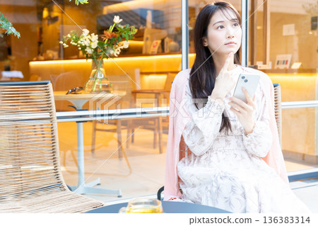 一位女士坐在咖啡館的露台座位上看著她的智慧型手機。 一位女士坐在咖啡館的露台座位上看著她的智慧型手機。 136383314