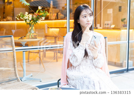 一位女士坐在咖啡館的露台座位上看著她的智慧型手機。 一位女士坐在咖啡館的露台座位上看著她的智慧型手機。 136383315