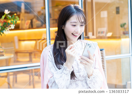 一位女士坐在咖啡館的露台座位上看著她的智慧型手機。 一位女士坐在咖啡館的露台座位上看著她的智慧型手機。 136383317