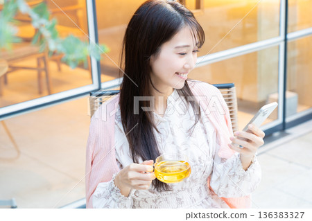 一位女士坐在咖啡館的露台座位上看著她的智慧型手機。 一位女士坐在咖啡館的露台座位上看著她的智慧型手機。 136383327