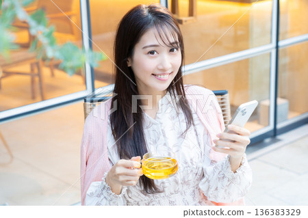 一位女士坐在咖啡館的露台座位上看著她的智慧型手機。 一位女士坐在咖啡館的露台座位上看著她的智慧型手機。 136383329
