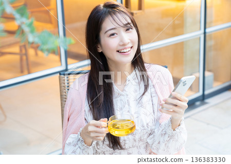 一位女士坐在咖啡館的露台座位上看著她的智慧型手機。 一位女士坐在咖啡館的露台座位上看著她的智慧型手機。 136383330