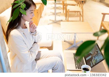 A young woman sitting on a sofa in a cafe and having a remote meeting on a computer A young woman sitting on a sofa in a cafe and having a remote meeting on a computer 136383342