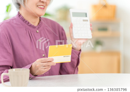 Smiling senior woman looking at her savings passbook in the living room of her home (retirement funds, asset management, deposits, income, living expenses) Smiling senior woman looking at her savings passbook in the living room of her home (retirement funds, asset management, deposits, income, living expenses) 136385796