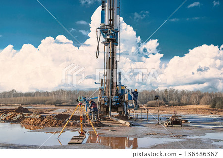 A drilling rig is actively working to reach oil and gas deposits in a rural area. Workers are present around the rig to monitor operations during the day A drilling rig is actively working to reach oil and gas deposits in a rural area. Workers are present around the rig to monitor operations during the day 136386367