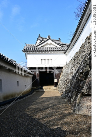 一座日本城堡在藍天的襯托下熠熠生輝:潔白的灰泥牆和通往雄偉的櫓門的道路。 一座日本城堡在藍天的襯托下熠熠生輝:潔白的灰泥牆和通往雄偉的櫓門的道路。 136386581