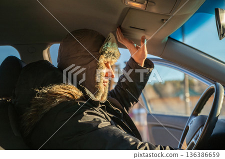 Man driver in a warm fur hat blocking sun with visor in car. Warming up the car engine before driving. Winter driving and travel concept. Cold weather journey. 136386659