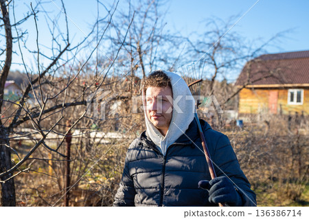 young Caucasian male farm worker with rake on a sunny autumn day in orchard. Farm work, harvest, cleaning, or gardening concept. Landowner, farmer, organic farm worker 136386714