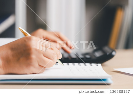 Hand writing notes in a notebook beside a calculator on office desk for budgeting and accounting. Personal finance planning, expense tracking, tax calculation, soft focus background, copy space. Hand writing notes in a notebook beside a calculator on office desk for budgeting and accounting. Personal finance planning, expense tracking, tax calculation, soft focus background, copy space. 136388137
