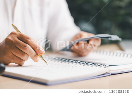 Hand writing notes in a notebook beside a calculator on office desk for budgeting and accounting. Personal finance planning, expense tracking, tax calculation, soft focus background, copy space. Hand writing notes in a notebook beside a calculator on office desk for budgeting and accounting. Personal finance planning, expense tracking, tax calculation, soft focus background, copy space. 136388138