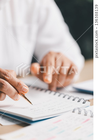 Hand writing notes in a notebook beside a calculator on office desk for budgeting and accounting. Personal finance planning, expense tracking, tax calculation, soft focus background, copy space. Hand writing notes in a notebook beside a calculator on office desk for budgeting and accounting. Personal finance planning, expense tracking, tax calculation, soft focus background, copy space. 136388139