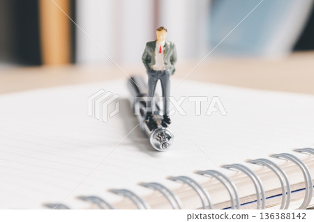 Miniature businessman standing on a pen over a spiral notebook, symbolizing leadership and planning. Business strategy, meeting notes, office productivity, consulting concept with copy space. 136388142