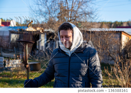 Smiling young white man, agricultural worker with rake on sunny autumn day on organic farm. Portrait from the waist up. Farm work, harvest, cleaning, gardening 136388161