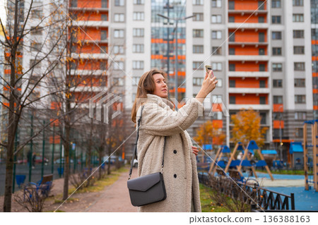 Caucasian European Woman takes photo with smartphone, writes message on social media and surfs news in residential area. Mobile internet problem and bad connection. Caucasian European Woman takes photo with smartphone, writes message on social media and surfs news in residential area. Mobile internet problem and bad connection. 136388163