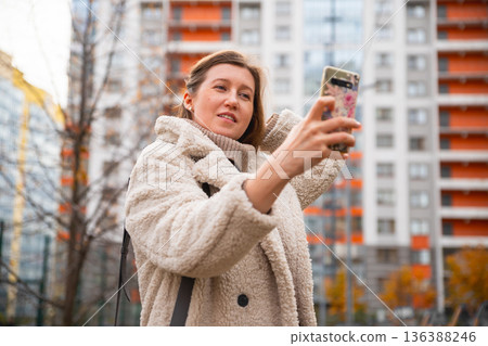 Making selfie woman on smartphone outdoors in an urban area with modern buildings. Young smiling European white female is taking selfie portrait with mobile phone. Making selfie woman on smartphone outdoors in an urban area with modern buildings. Young smiling European white female is taking selfie portrait with mobile phone. 136388246