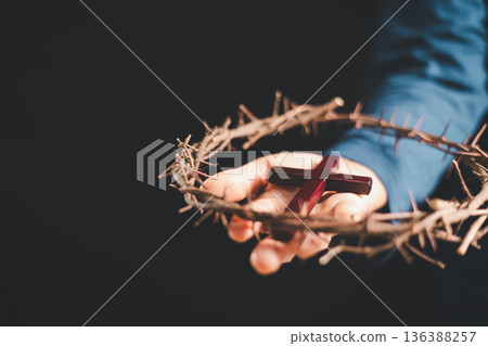 Hand placing a wooden cross above a crown of thorns on an open Bible, dark background and dramatic light with copy space, symbolizing crucifixion, sacrifice, Holy Week and Easter faith. 136388257