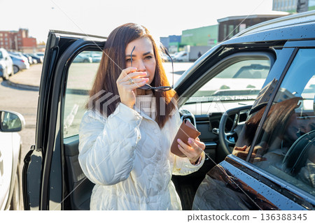 Woman standing by car door, holding sunglasses and wallet. Lifestyle scene of a person getting out of a vehicle in a parking lot. 136388345