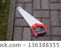 Hand saw with red and black handle on a brick path, detail of a sharp blade and teeth for cutting, gardening tool. 136388355